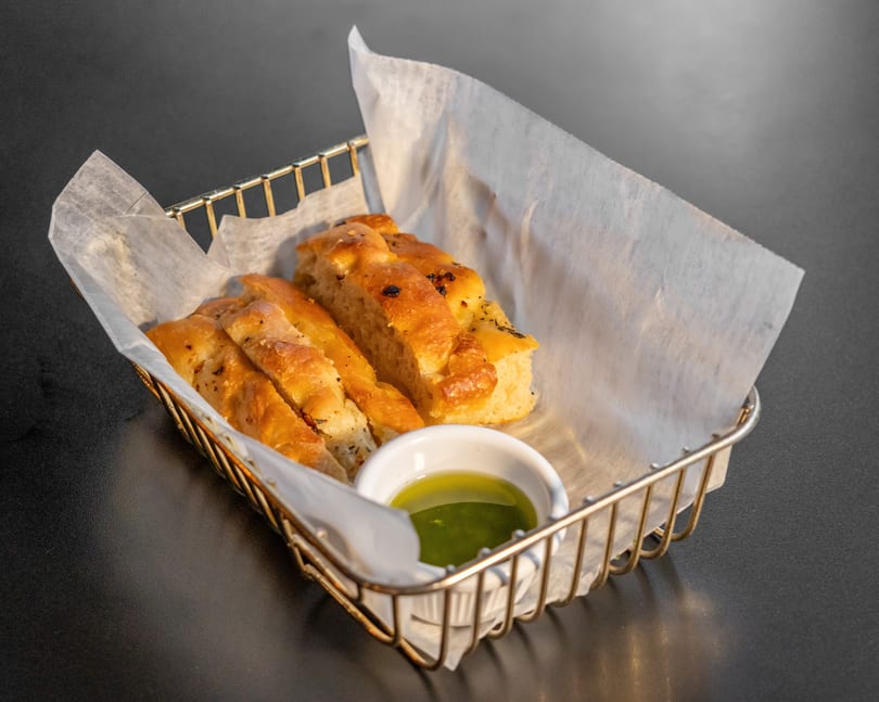 Golden focaccia bread slices in a wire basket with parchment paper and a small dish of olive oil.