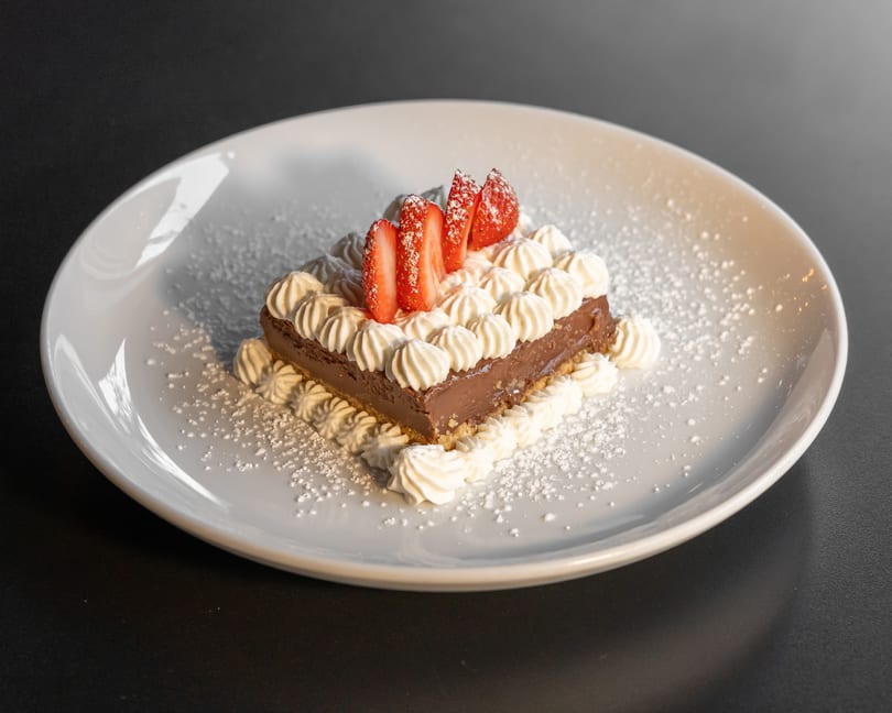 Chocolate tart with whipped cream and sliced strawberries, dusted with powdered sugar on a white plate.