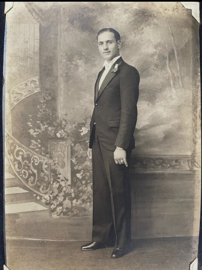 A vintage black and white portrait of a man in a tuxedo standing next to a painted staircase.