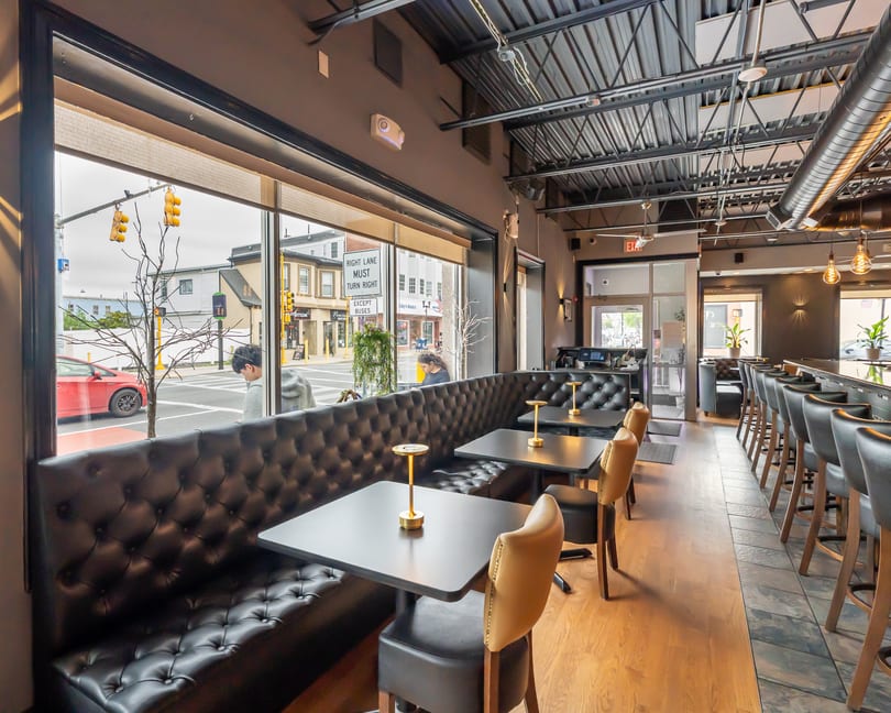 Modern restaurant interior with black tufted booths, small tables, and a long bar with stools.