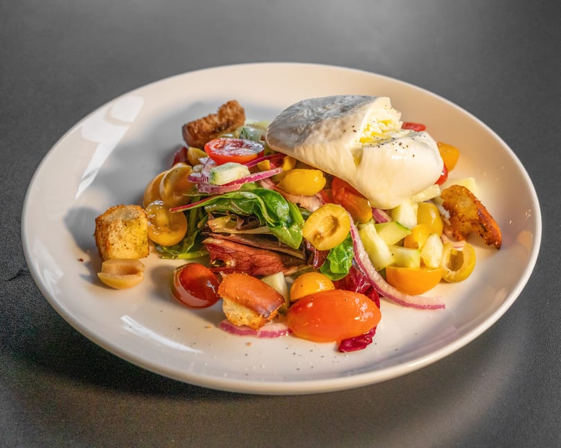 A vibrant salad with burrata cheese, cherry tomatoes, red onion, olives, and croutons on a white plate.