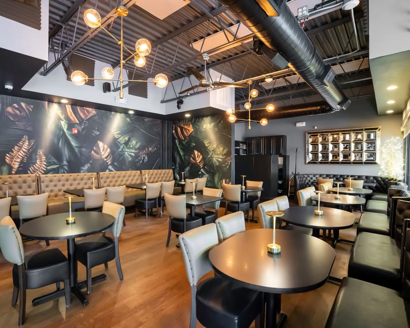 Modern restaurant interior with dark tables, light chairs, banquette seating, and a tropical leaf mural.