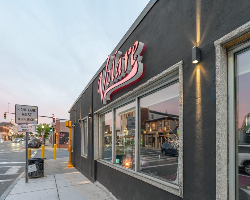 Exterior of Volare restaurant at dusk, with a glowing red sign and street reflections in the windows.