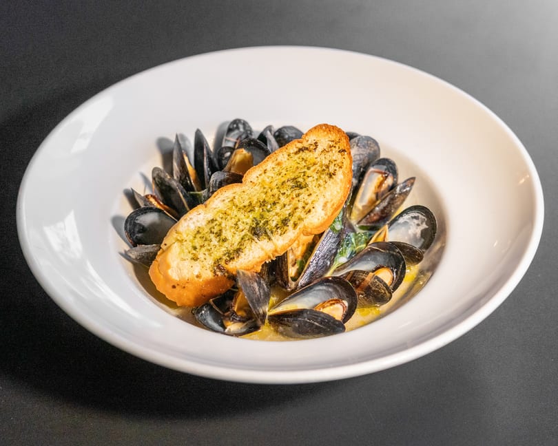 Steamed mussels in a white bowl with broth and a slice of garlic bread on top.