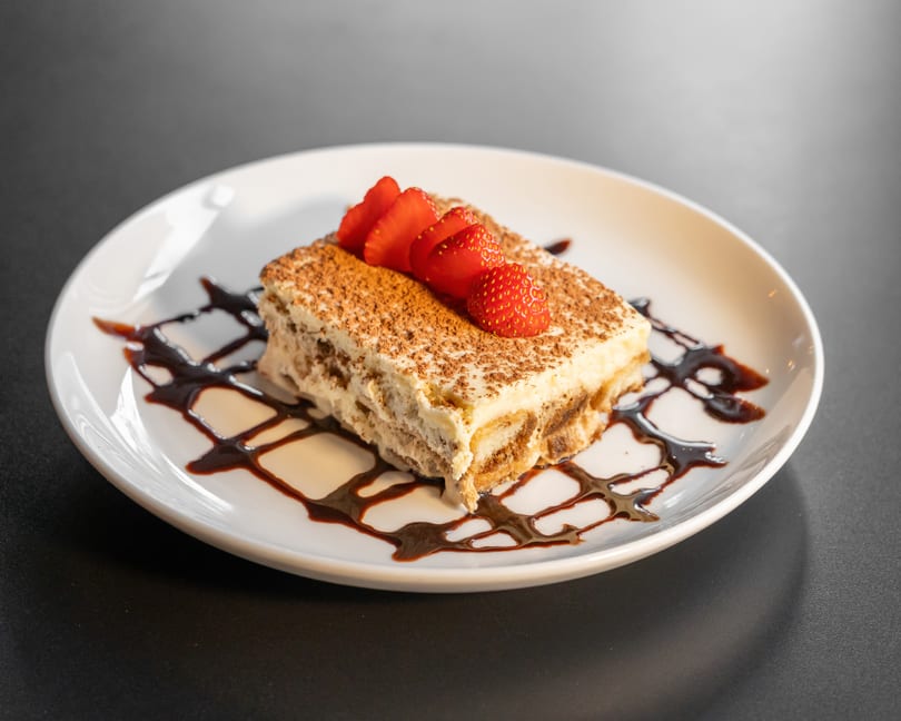 A slice of tiramisu with cocoa powder, fresh strawberries, and chocolate drizzle on a white plate.