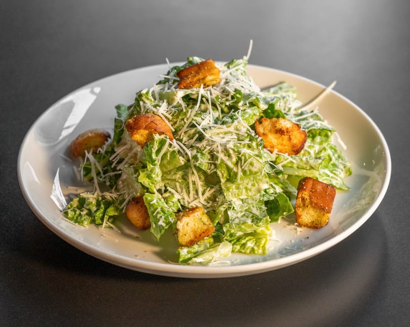 A Caesar salad with romaine lettuce, croutons, and shredded Parmesan cheese on a white plate.